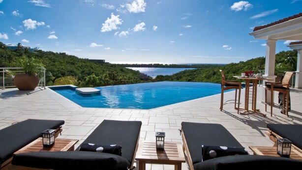 A pool area with lounge chairs and a table, surrounded by lush greenery and a view of a lake.