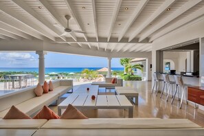 A spacious indoor area with a ceiling fan, a seating area with sofas and a table, and a view of the ocean. - Sandyline (4/7 Rooms) (Les Terres Basses)
