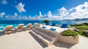 Uma área de estar ao ar livre com luz do sol, cadeiras de lounge e um sofá, com vista para um mar azul claro e montanhas ao longe.