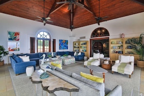 A spacious living room with a high ceiling, a large window, and a fireplace. There are multiple seating areas with sofas and armchairs.