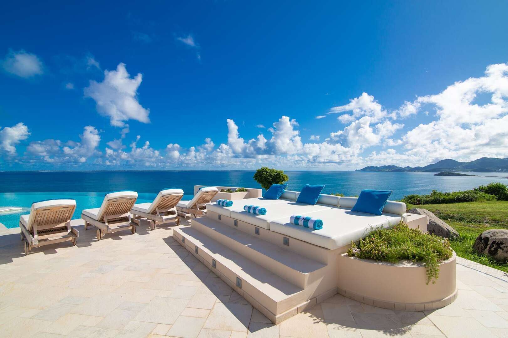 A sunlit outdoor seating area with lounge chairs and a sofa, overlooking a clear blue sea and distant mountains.
