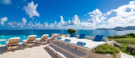 A sunlit outdoor seating area with lounge chairs and a sofa, overlooking a clear blue sea and distant mountains.