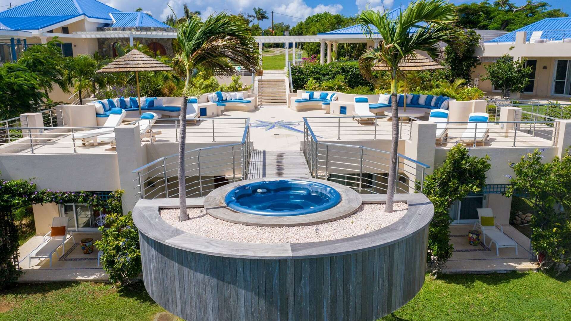 Uma banheira de hidromassagem circular rodeada por um deck com espreguiçadeiras, uma escadaria que leva a uma área coberta e uma piscina com teto azul.