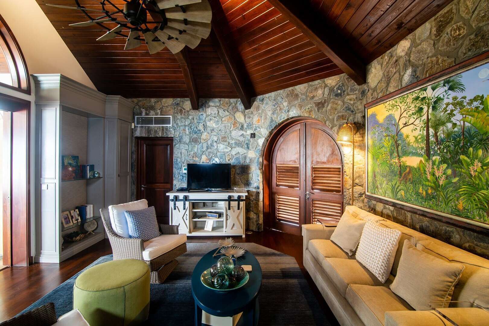 A cozy living room with a stone wall, wooden ceiling, and a large painting of a tropical scene.