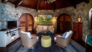 Um quarto aconchegante com paredes de pedra, vigas de madeira no teto, um grande ventilador de teto, televisão, um sofá, duas cadeiras de vime, uma mesa de centro e um armário.