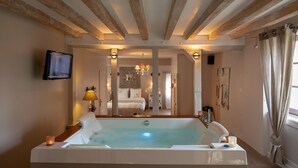A spacious room with a large bathtub, wooden beams on the ceiling, a television on the wall, and a cozy bed in the background. - Romantic suite with private jacuzzi 1 hour from Paris (Vendières)