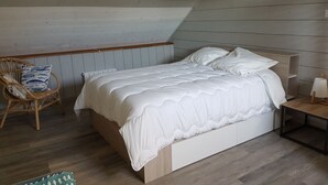 Quarto com teto de madeira inclinado, cama com roupa de cama branca, cadeira de vime com almofada e mesa de cabeceira com abajur.