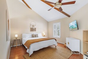 Une chambre à coucher avec un lit, des lampes de chevet, un ventilateur de plafond, une fenêtre, un téléviseur fixé au mur et une commode blanche.