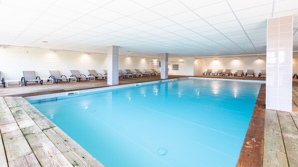 A large indoor swimming pool with wooden deck edges, multiple lounge chairs, and a tiled ceiling.