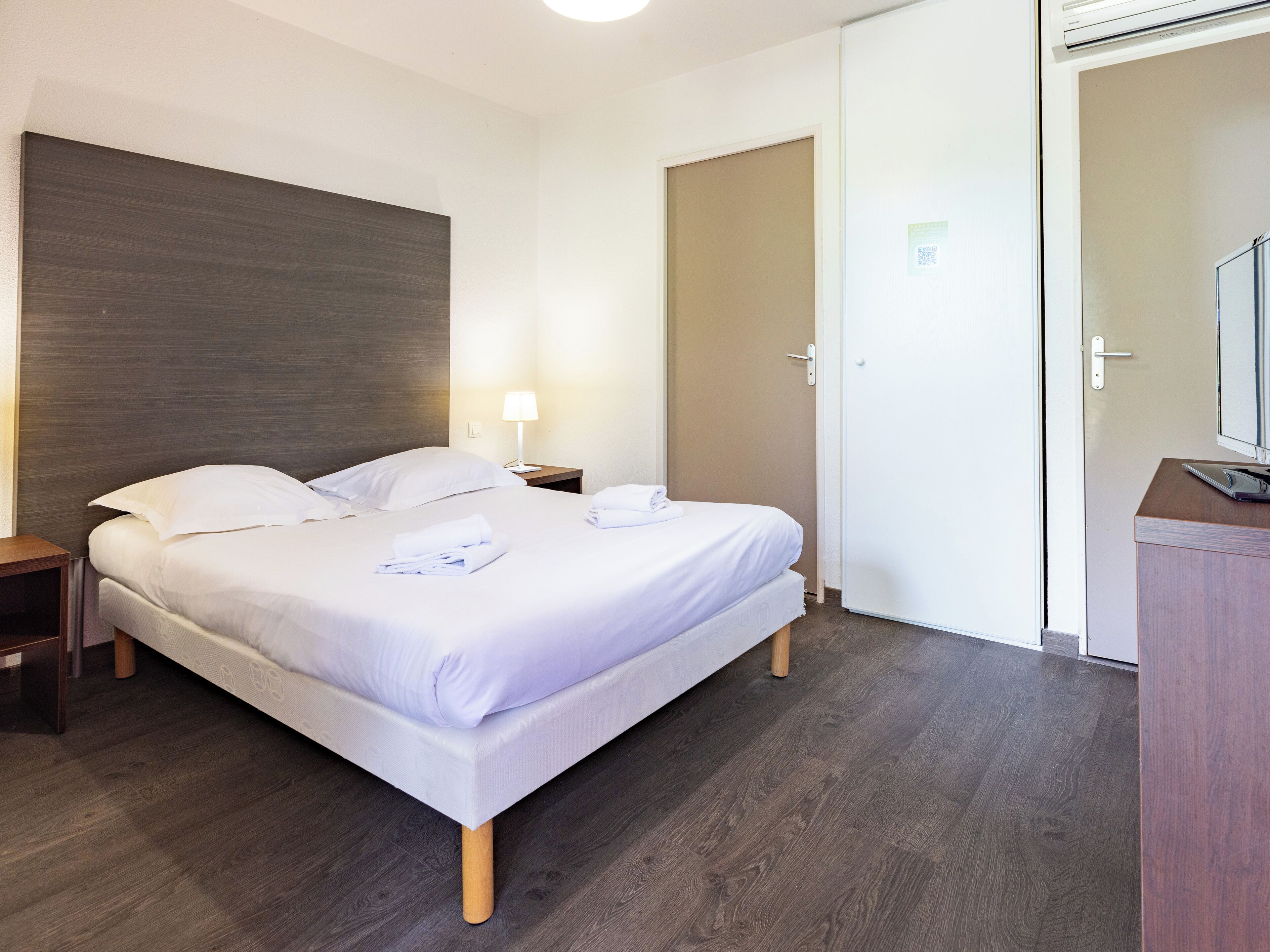 Una cama bien hecha con sábanas blancas, una lámpara de noche y una mesita de noche de madera.