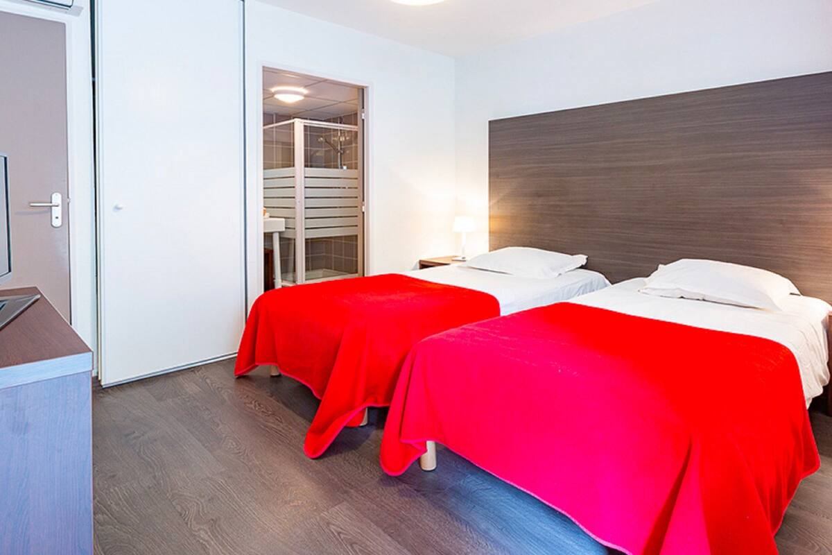 Ein Hotelzimmer mit zwei Betten, jedes mit roter Bettdecke, einem Holz-Kopfteil und einem Nachttisch mit Lampe.