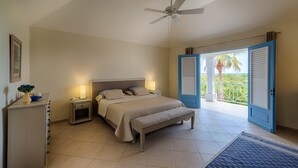 A bedroom with a bed, bedside lamps, a nightstand, a bench, a ceiling fan, and a view through an open door. - Oceane (2/4 Rooms) Luxury villa (Marigot)