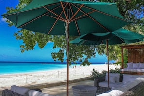 Eine Strandlounge mit einem großen, grünen Sonnenschirm, Korbmöbeln und Blick auf das Meer.
