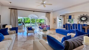 A spacious living room with a glass coffee table, beige sofas with blue cushions, and a view of a lush garden through large windows. - Moon Dance Luxury villa (Sandy Lane)