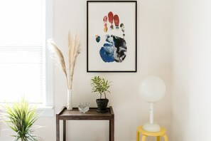Une œuvre d’art encadrée représentant une empreinte de main sur un mur, une petite table avec une plante en pot, un vase contenant des plantes séchées et une lampe sur pied.