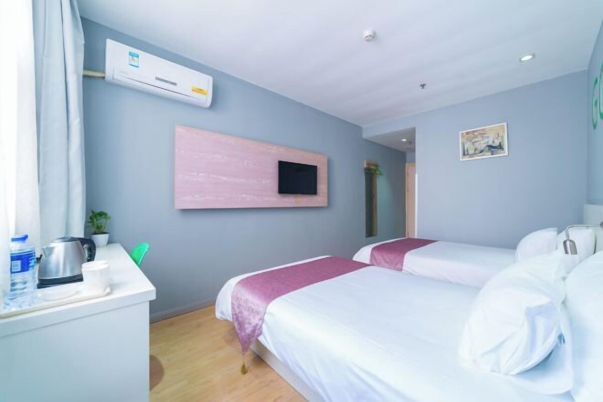 A hotel room with two beds, a desk with a kettle and a cup, an air conditioning unit, and a mounted TV.