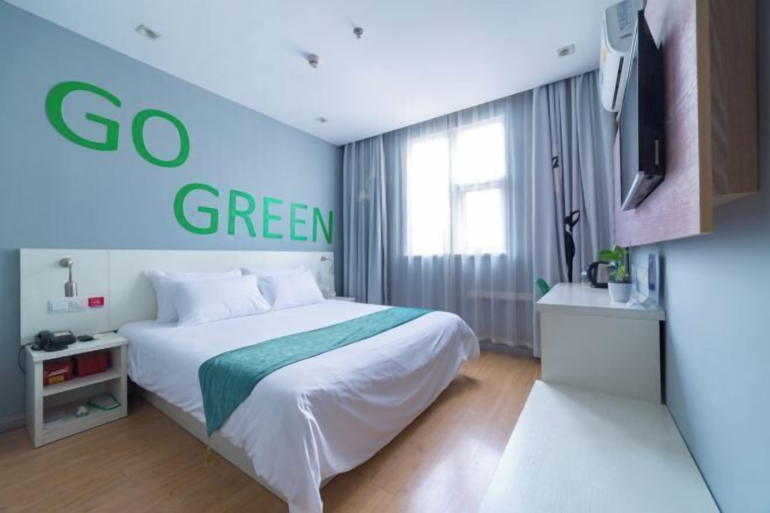 A hotel room with a bed, a bedside table, a TV, and a wall with 'GO GREEN' text.