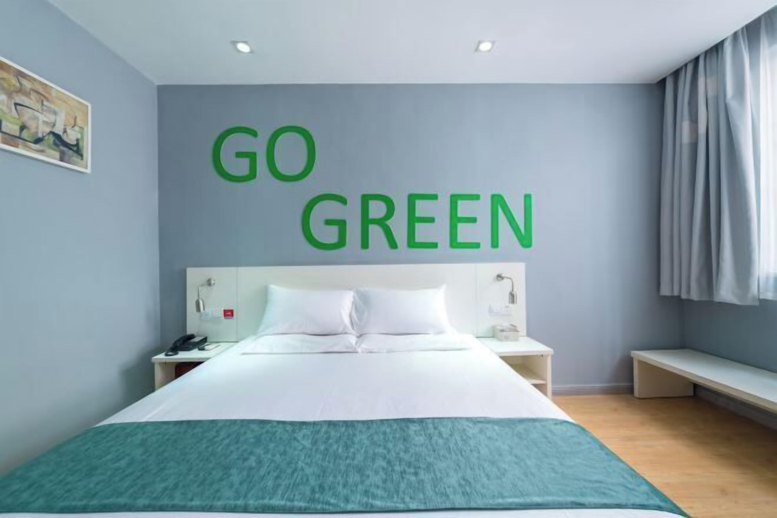 A hotel room with a bed, a nightstand, a bedside table, a bench, and a wall with the text 'GO GREEN'.