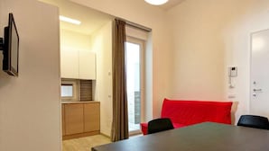 A compact room with a dining table, a red couch, and a kitchen area visible through an open door. - Appartamento Ariel   La Perla del Mare di Polignano (Polignano a Mare)