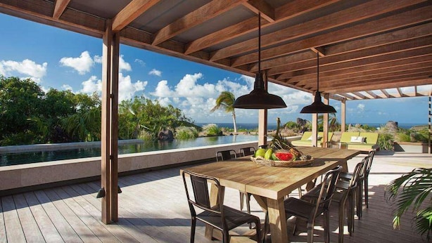 Un área de comedor al aire libre con una mesa y sillas de madera, con vista a una piscina y exuberante vegetación.