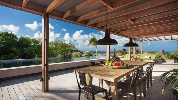 Un área de comedor al aire libre con una mesa y sillas de madera, con vista a una piscina y exuberante vegetación.