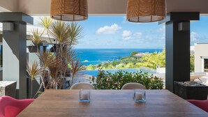 A dining area with a view of the ocean, featuring a wooden table, chairs, and two candle holders. - Vague Bleue (1/3 Rooms) (Lurin)