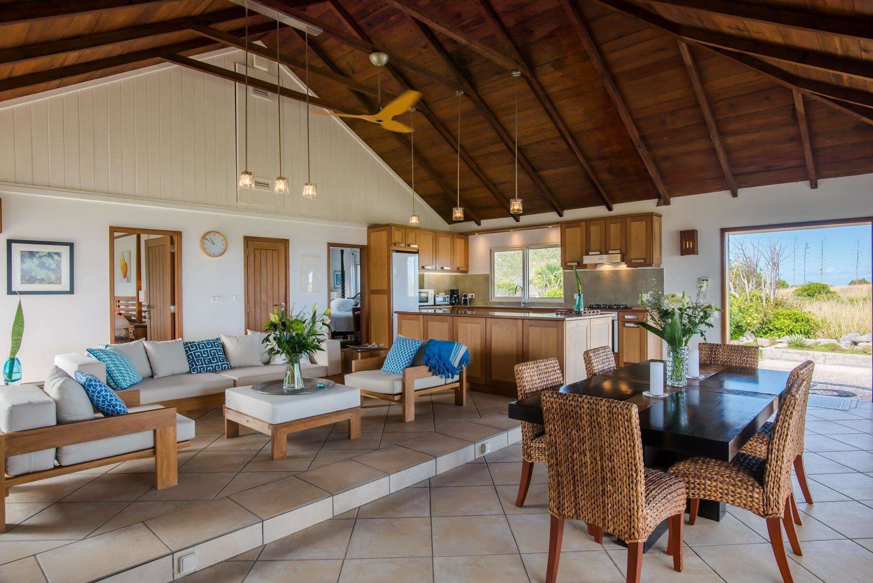A spacious living area with a kitchen and dining area, featuring wooden furniture, a ceiling fan, and large windows.