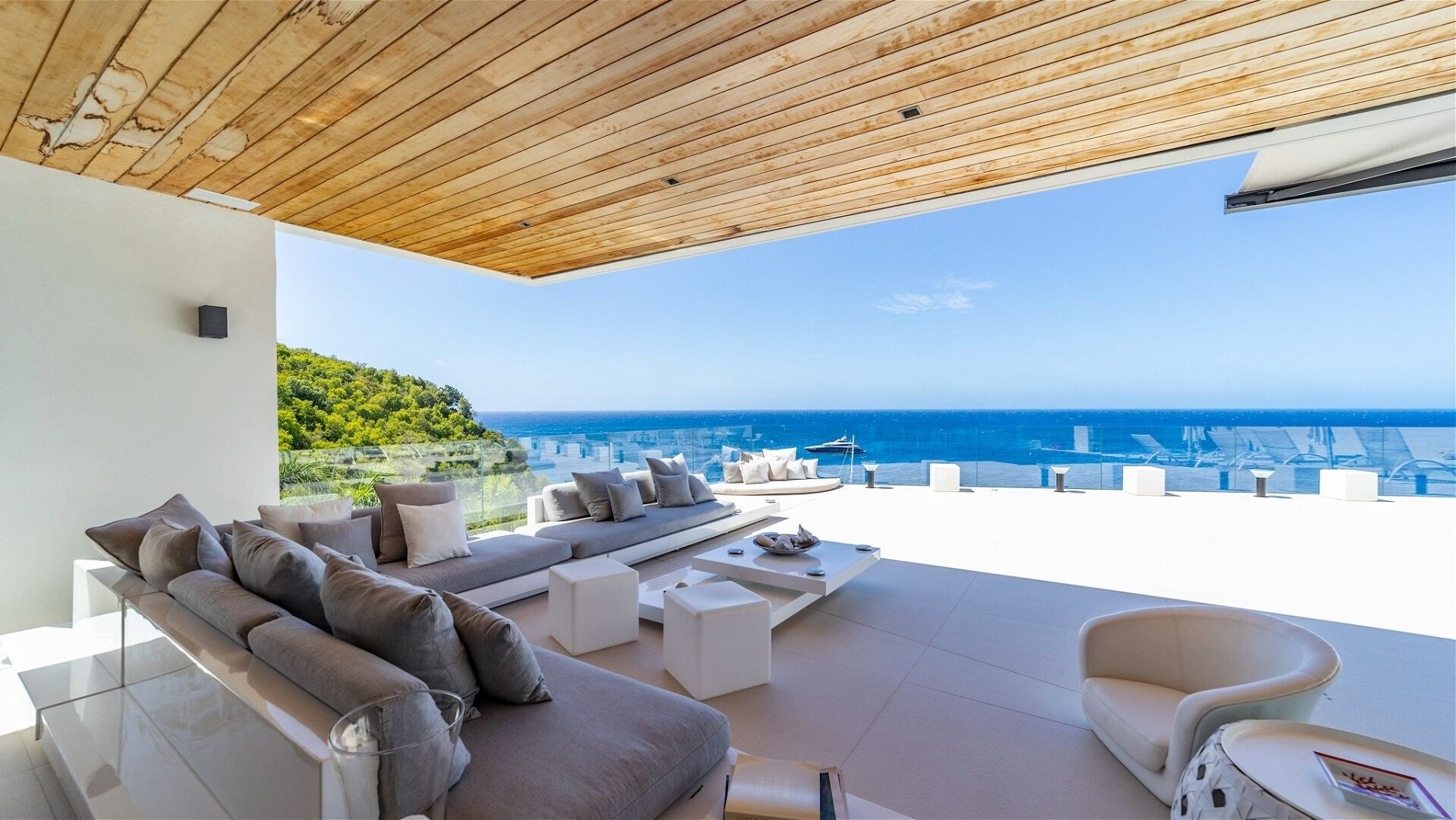 A modern outdoor living area with a wooden ceiling, white furniture, and a view of the sea.