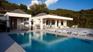 Un espace moderne aménagé au bord de la piscine, avec une piscine à l’eau d’un bleu limpide, des chaises longues blanches et un coin salon couvert équipé de tables et de chaises.