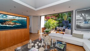 Un salon moderne doté d’un grand téléviseur, d’un canapé blanc, d’une table basse agrémentée d’objets décoratifs et offrant une vue sur un jardin tropical grâce à de grandes fenêtres.