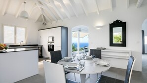 A modern kitchen with white cabinetry, a dining area with a round table, and a view of the ocean. - Taniko (1/3 Rooms) Luxury villa (Colombier)