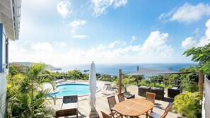 A pool area with a view of the ocean, outdoor furniture, and lush greenery.