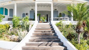 Una escalera conduce a un área exterior cubierta con una mesa y sillas, rodeada de exuberante follaje y plantas.