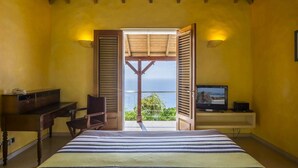 Quarto com cama, escrivaninha, cadeira, televisão e vista para o mar através de persianas abertas.