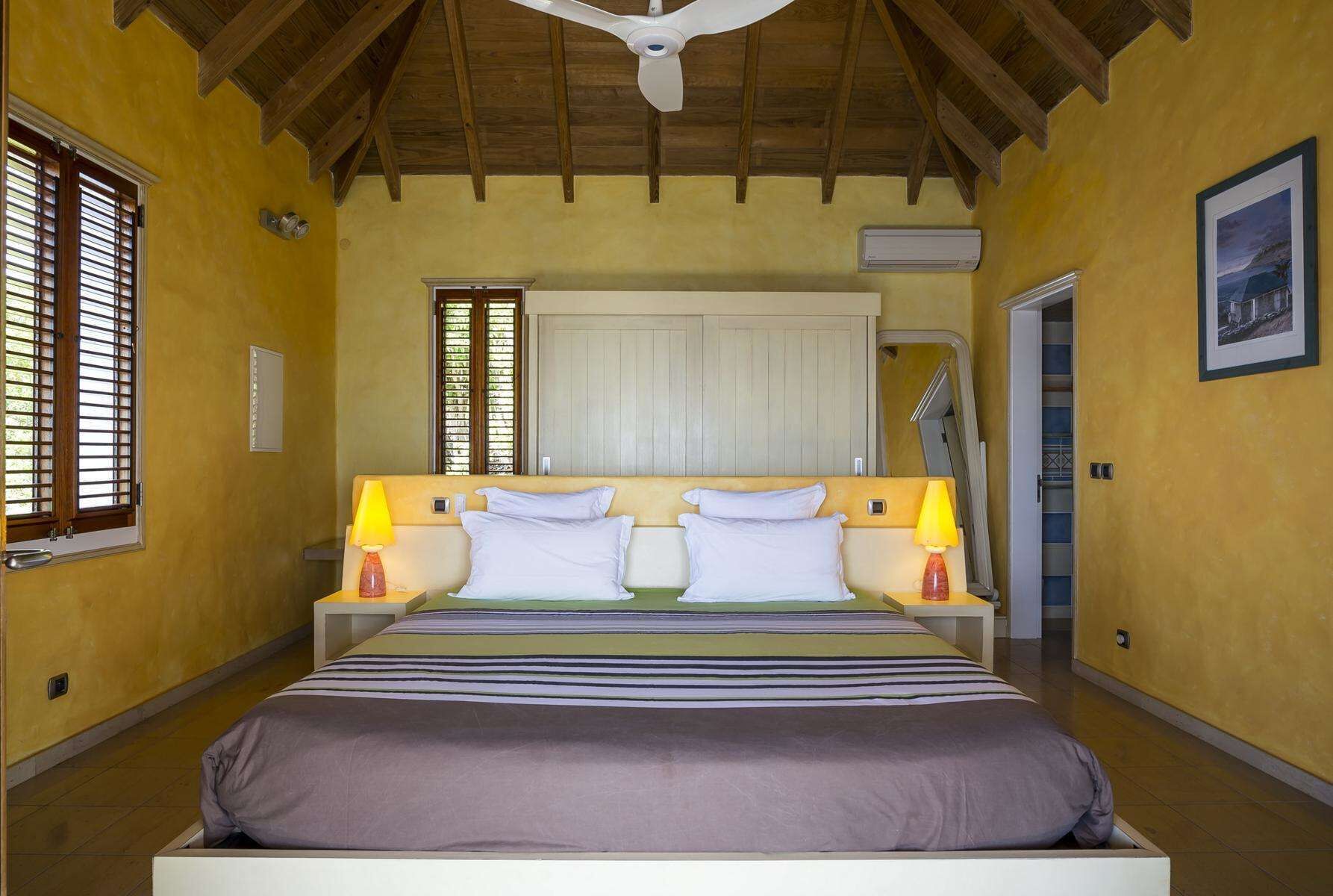 Un dormitorio con una cama, dos lámparas de noche, una ventana con persianas, un ventilador de techo y una puerta que da a otra habitación.