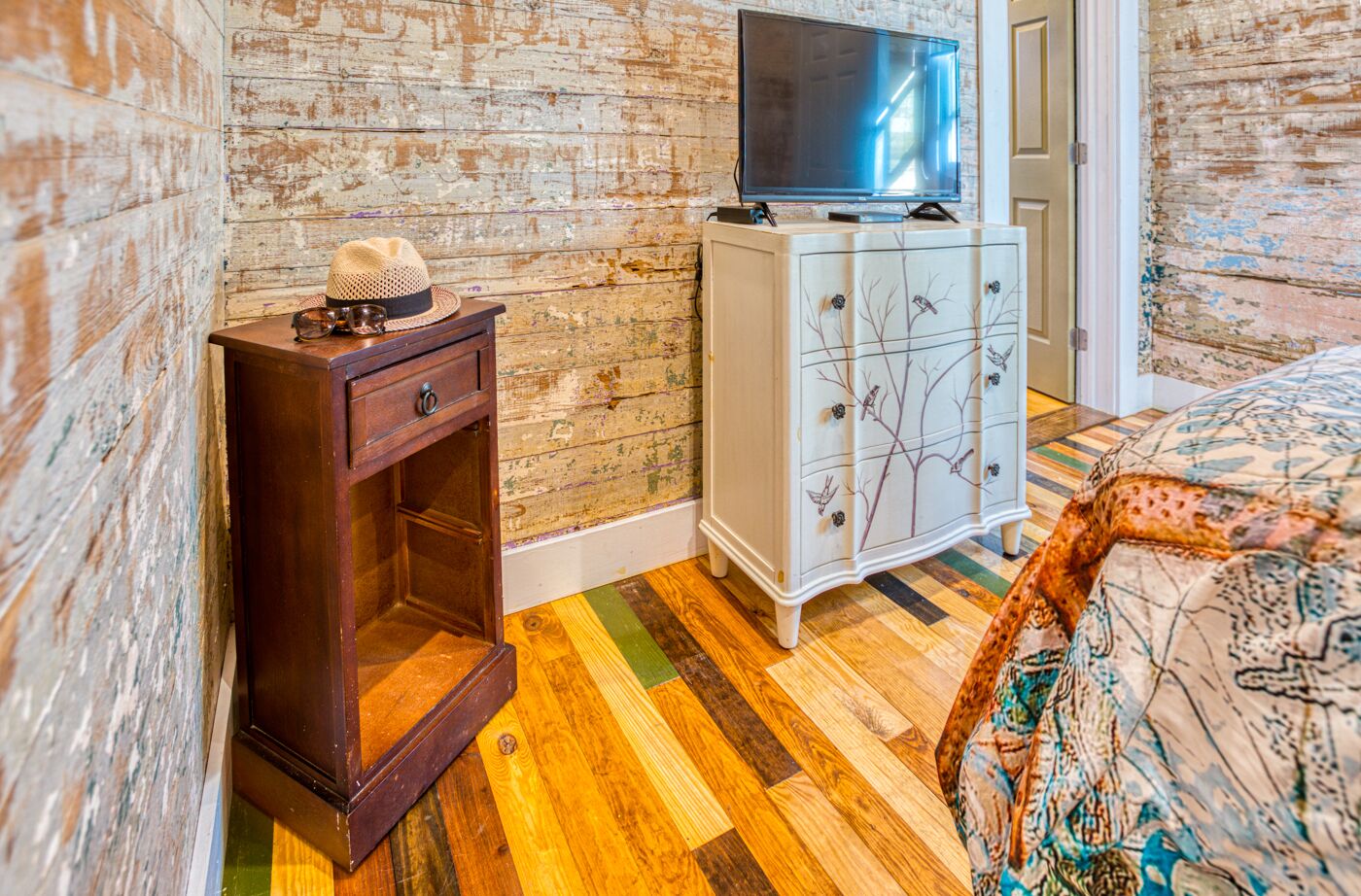 A room with a wooden floor, a TV on a cabinet, a small wooden cabinet, and a bed with a patterned bedspread.