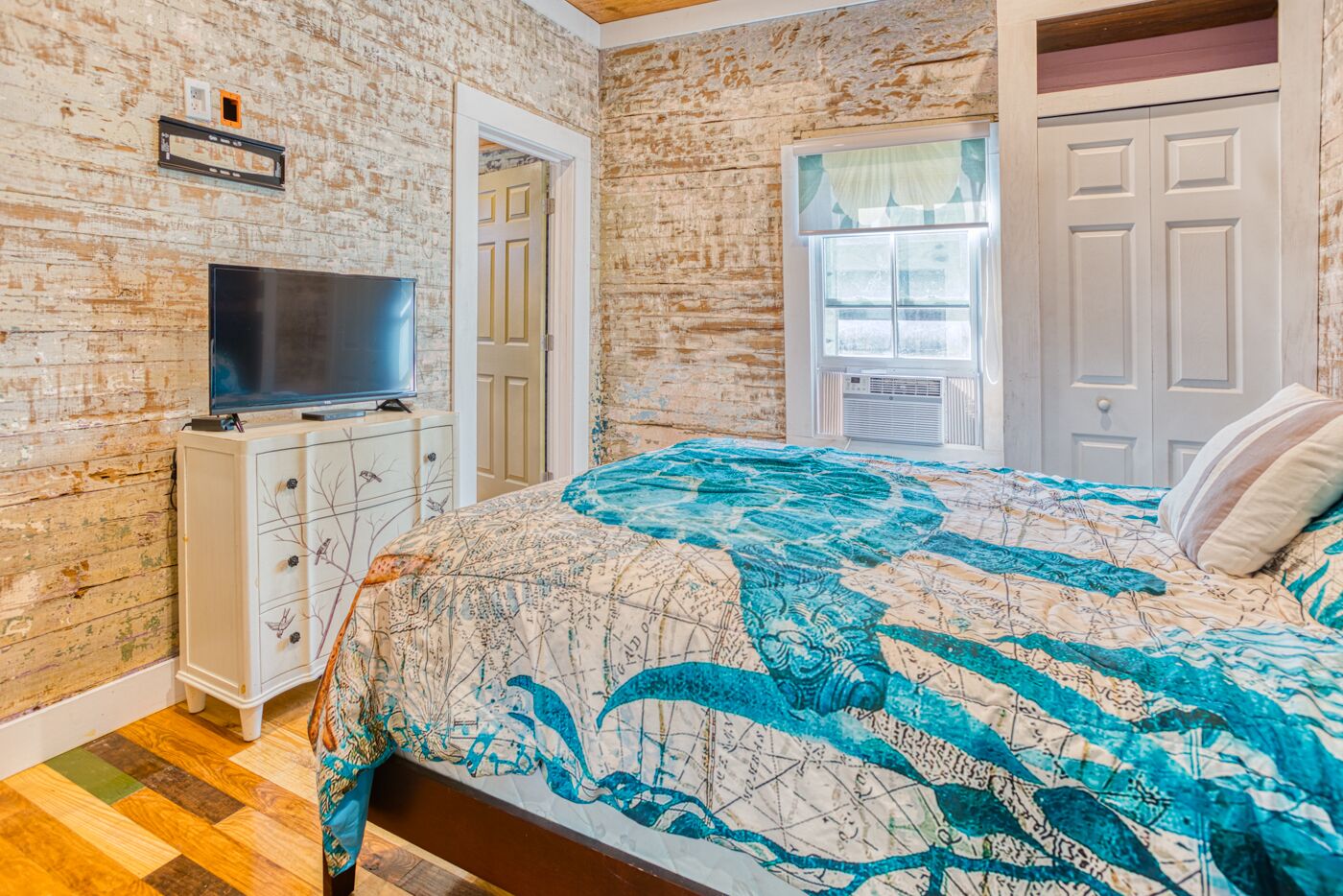 A bedroom with a brick wall, a bed with a map-themed bedspread, a TV on a dresser, and a window with a view.