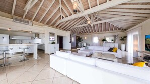 Un salón comedor amplio con techo alto y vigas a la vista, una cocina moderna con armarios blancos, y un área de comedor con una mesa y sillas blancas.