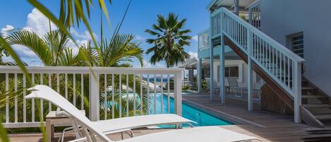A pool area with two lounge chairs, a wooden deck, a white railing, and a staircase leading to a building.