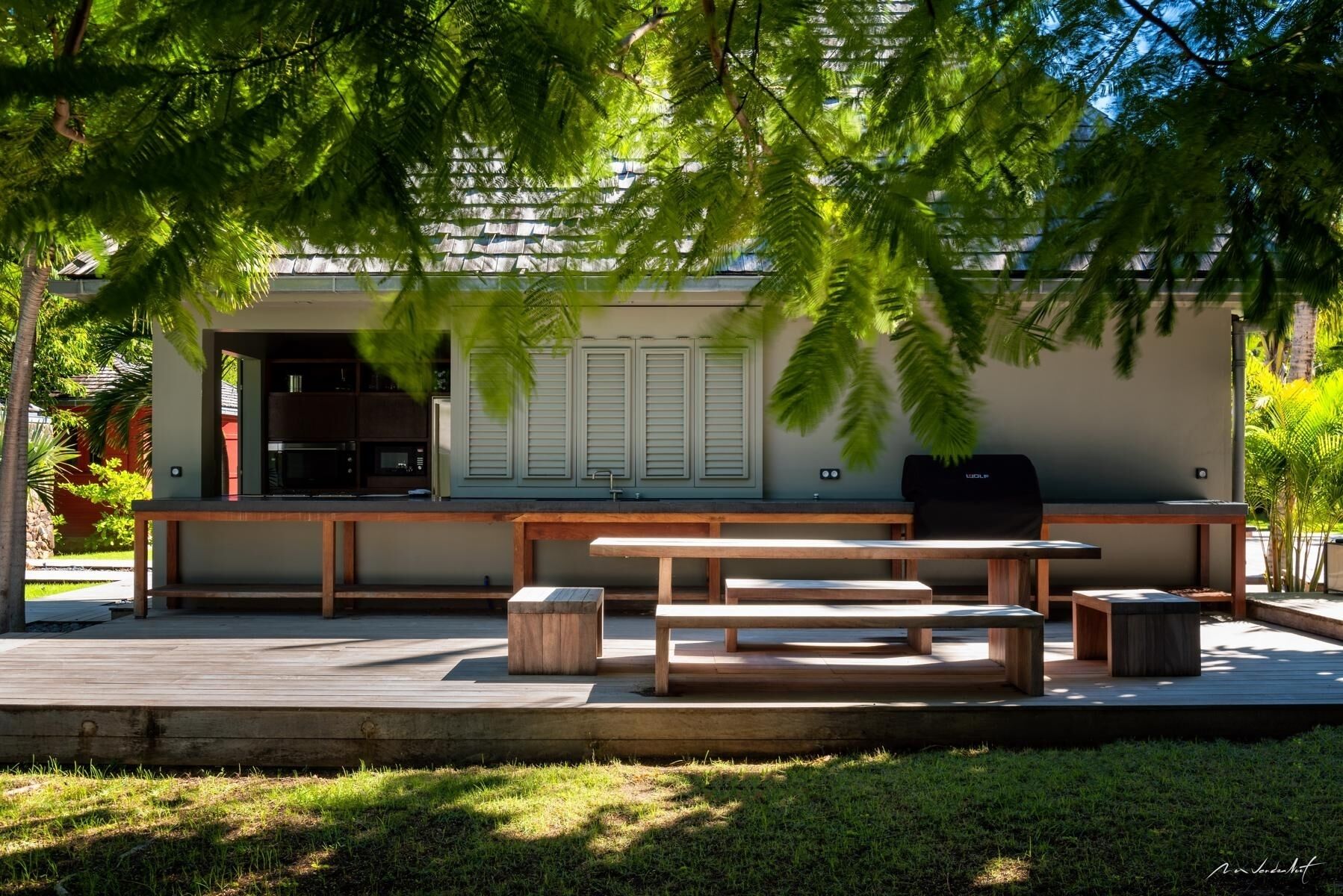 Eine Außenpatio mit Holzbänken und einem Grill, umgeben von Grünpflanzen und einem Gebäude mit heller Fassade.