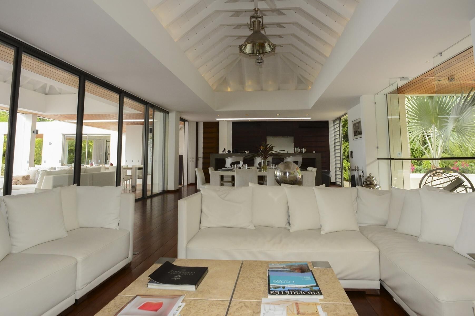 A spacious living area with a large white sofa, a coffee table with books, and a view of the kitchen and outdoor greenery.