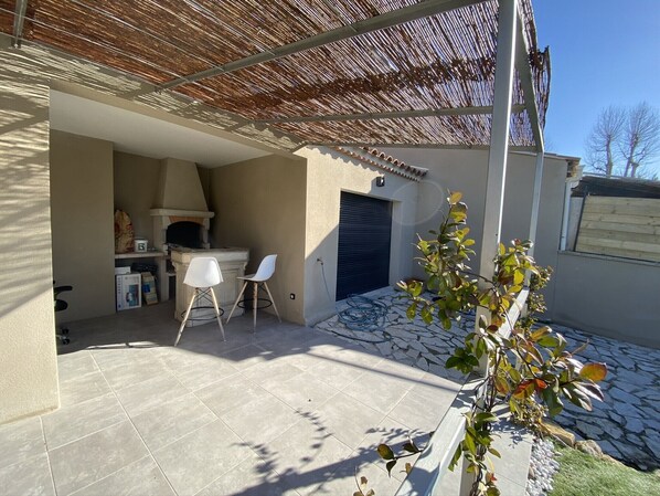 A patio area with a stone floor, two white chairs, a pizza oven, and a covered seating area with a wooden roof.