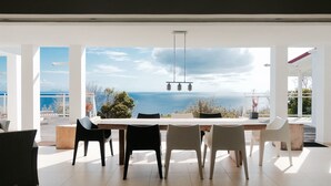Un espace repas offrant une vue sur la mer, avec une longue table et des chaises, un luminaire suspendu et un intérieur blanc.