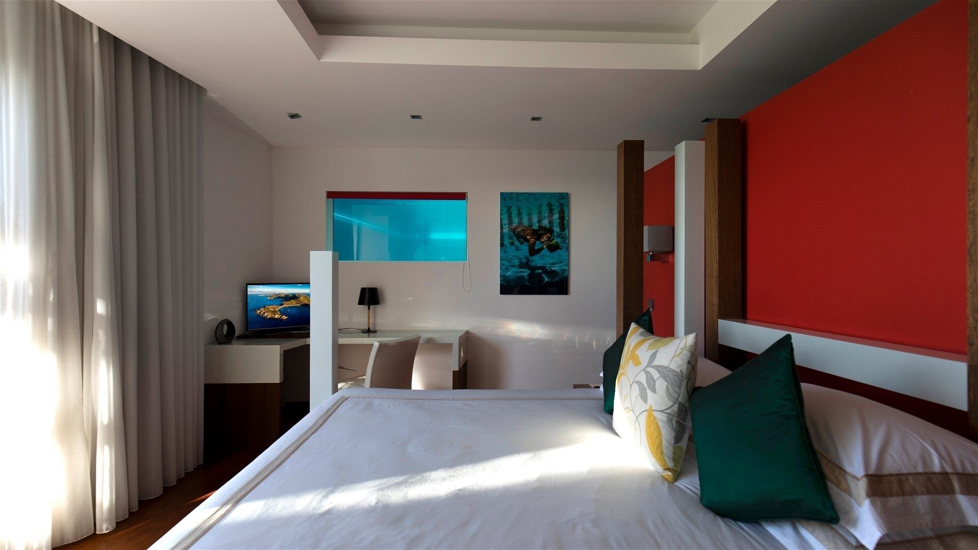 Habitació d'hotel amb un llit gran, un escriptori amb un ordinador, una finestra amb cortines i una paret vermella decorativa.