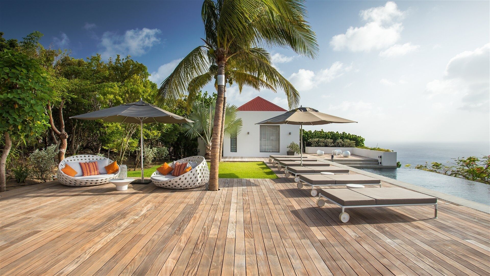 Een houten terras met lounge stoelen en parasols, een klein gebouw met een rood dak en uitzicht op de oceaan.