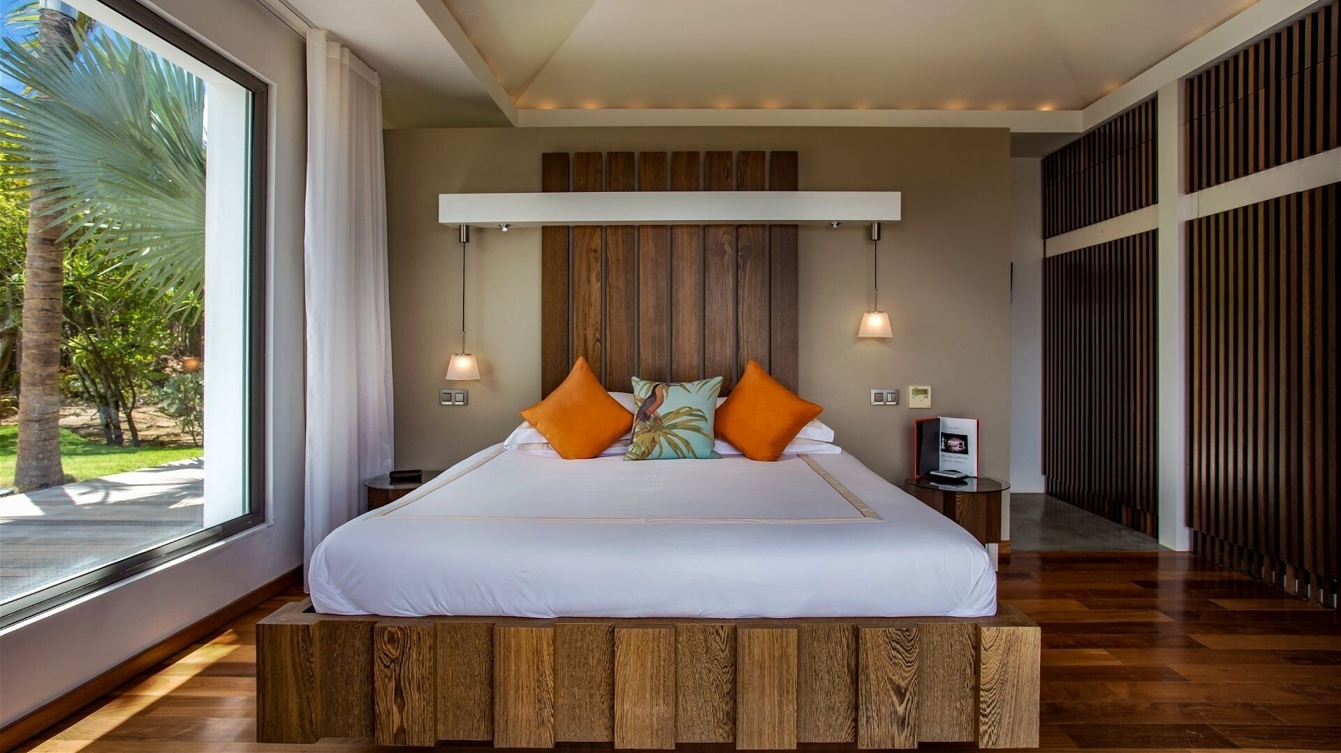 Ein modernes Schlafzimmer mit einem großen Bett, einem Holzkopfteil, zwei Nachttischlampen und Blick auf Palmen durch große Fenster.