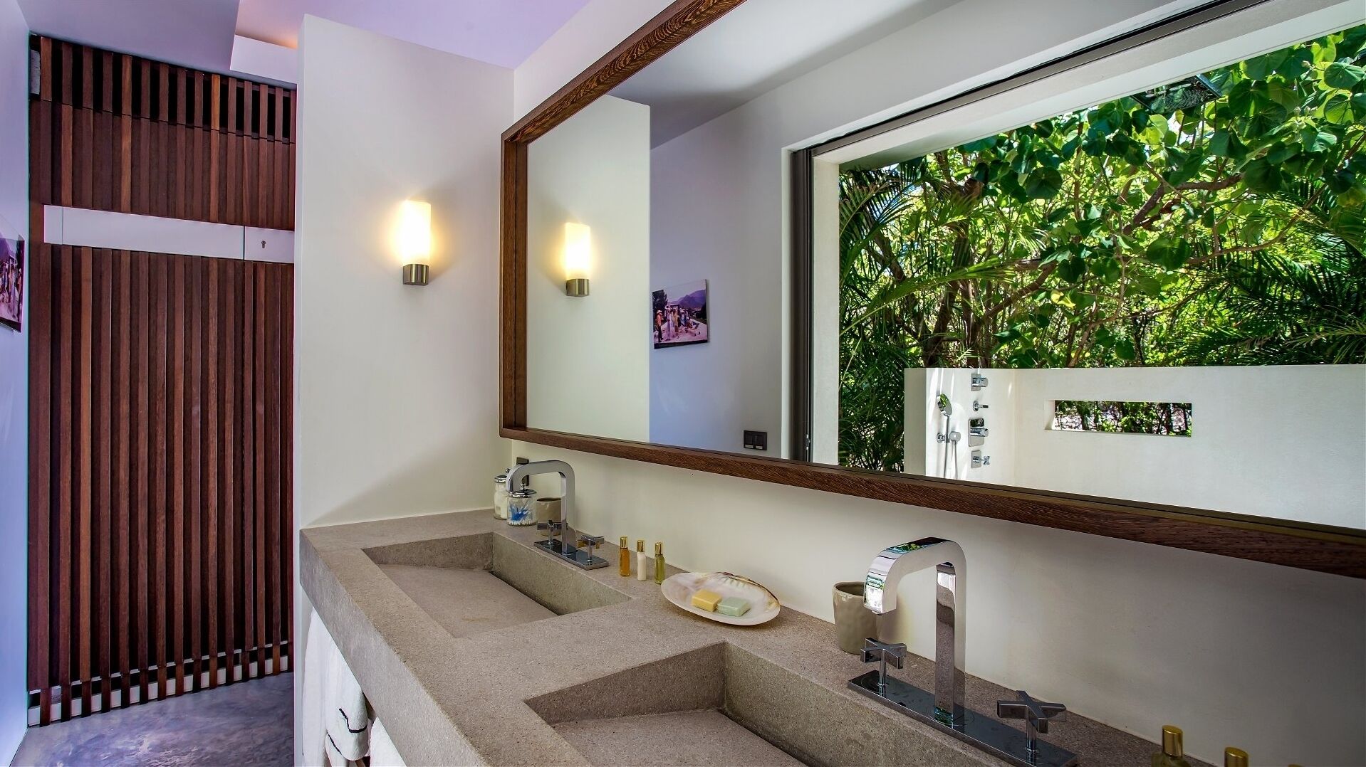 A bathroom with a double sink, a large mirror, and a view of a lush green garden.