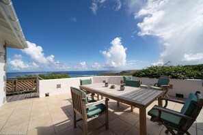 Een terras buiten met een houten tafel en stoelen, uitzicht op de oceaan en een betegelde vloer.