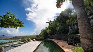 Ein Schwimmbecken mit Holzdeck und Steinfliesen, umgeben von üppiger Vegetation und Blick auf den Ozean.