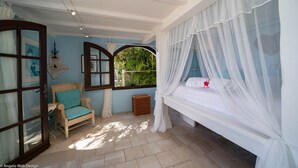 Ein Schlafzimmer mit einem Himmelsbett, einem Korbstuhl, einem kleinen Fenster mit Blick auf Grünflächen und einem an der Wand befestigten Regal.